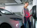 Kobieta ładuje biały samochód elektryczny. Czy od 2026 roku każda działka będzie musiała mieć przyłącze do ładowania aut elektrycznych?