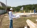 Inżynier z planami analizuje teren budowy, zastanawiając się, ile arów pod dom będzie potrzebne. Pracownicy w niebieskich kombinezonach kontynuują montaż ścian.