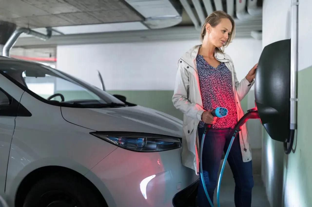 Kobieta ładuje biały samochód elektryczny. Czy od 2026 roku każda działka będzie musiała mieć przyłącze do ładowania aut elektrycznych?
