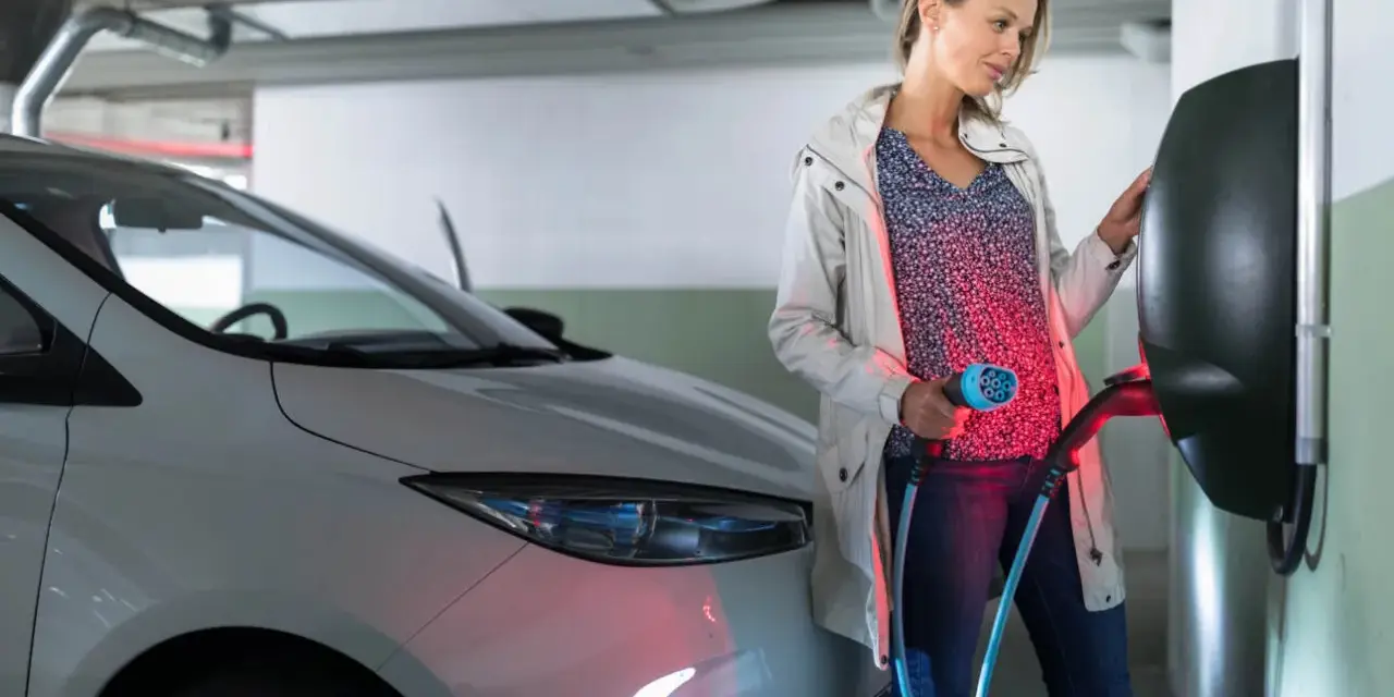 Kobieta ładuje biały samochód elektryczny. Czy od 2026 roku każda działka będzie musiała mieć przyłącze do ładowania aut elektrycznych?