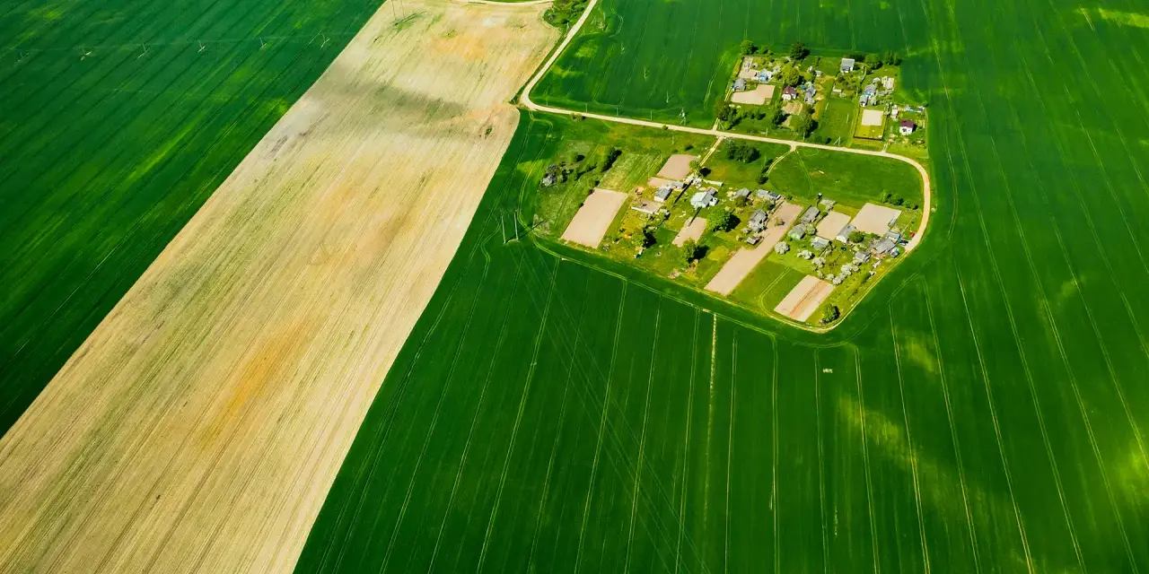 Widok z lotu ptaka na pola uprawne i wieś. Różne odcienie zieleni i żółcieni sugerują różne rodzaje gleby, co może pomóc w zrozumieniu, jak sprawdzić klasę gruntu.