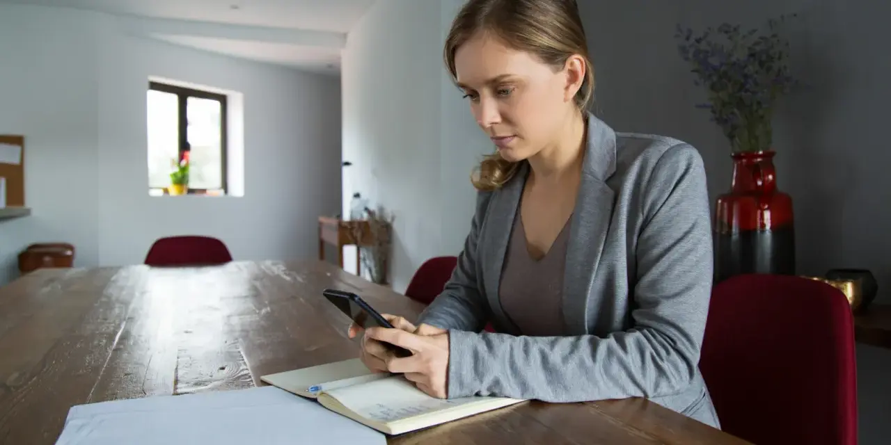 Kobieta z telefonem w ręku przy drewnianym stole. Analizuje dane, szukając odpowiedzi, dlaczego najem średnioterminowy staje się bezpieczniejszą alternatywą dla właścicieli.