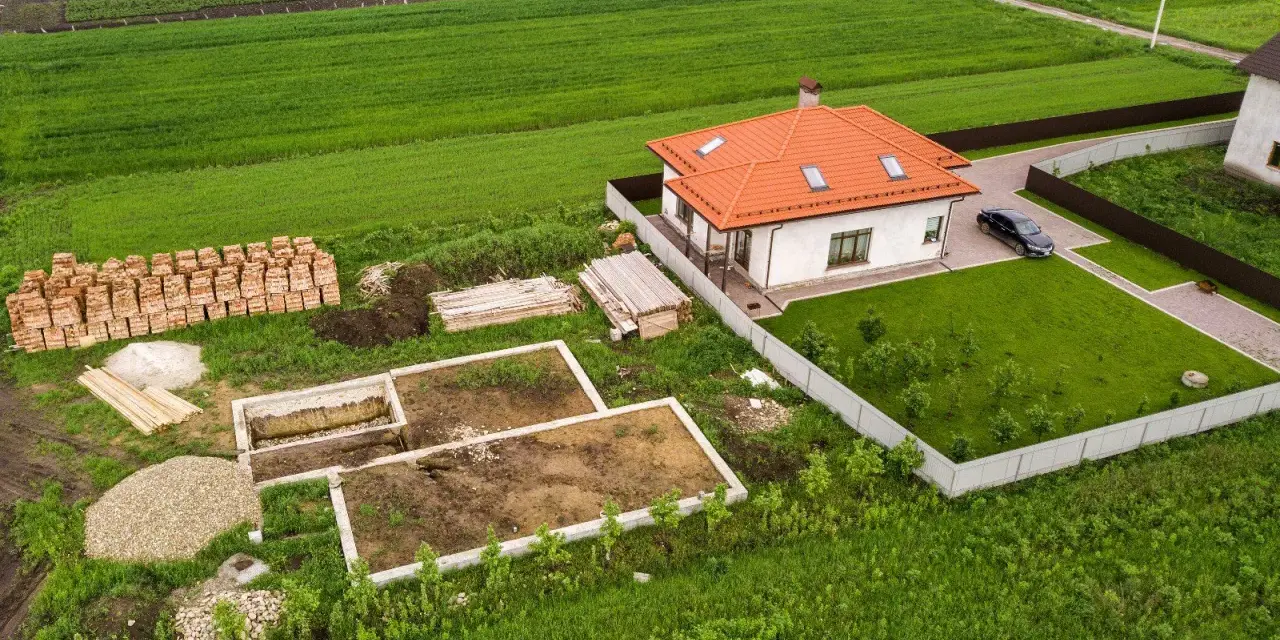 Budowa domu w toku: fundamenty, sterty cegieł i materiałów budowlanych. Obok gotowy dom i pole. Ile arów musi mieć działka budowlana?