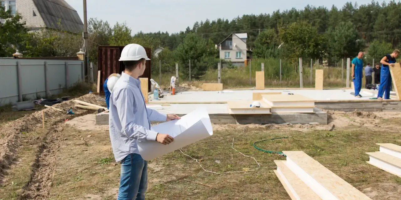 Inżynier z planami analizuje teren budowy, zastanawiając się, ile arów pod dom będzie potrzebne. Pracownicy w niebieskich kombinezonach kontynuują montaż ścian.