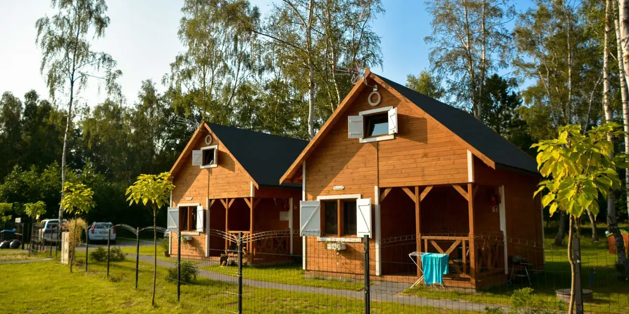 Dwa drewniane domki rekreacji indywidualnej, idealne na wynajem, stoją na zielonej trawie wśród drzew.