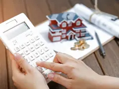 Dłoń na kalkulatorze, obok model domu, monety i plany. Wycena działki przez rzeczoznawcę będzie kluczowa dla banku od marca 2026.