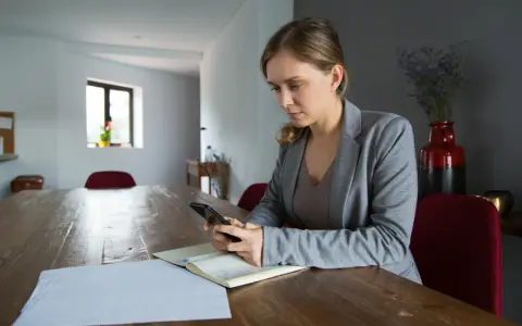 Kobieta z telefonem w ręku przy drewnianym stole. Analizuje dane, szukając odpowiedzi, dlaczego najem średnioterminowy staje się bezpieczniejszą alternatywą dla właścicieli.