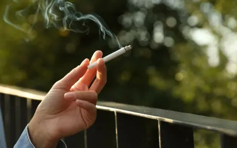 Dłoń trzyma zapalonego papierosa, z którego unosi się dym. Czy to prawda o rzekomym zakazie palenia papierosów na balkonach w nowych regulaminach?