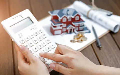 Dłoń na kalkulatorze, obok model domu, monety i plany. Wycena działki przez rzeczoznawcę będzie kluczowa dla banku od marca 2026.