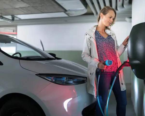 Kobieta ładuje biały samochód elektryczny. Czy od 2026 roku każda działka będzie musiała mieć przyłącze do ładowania aut elektrycznych?