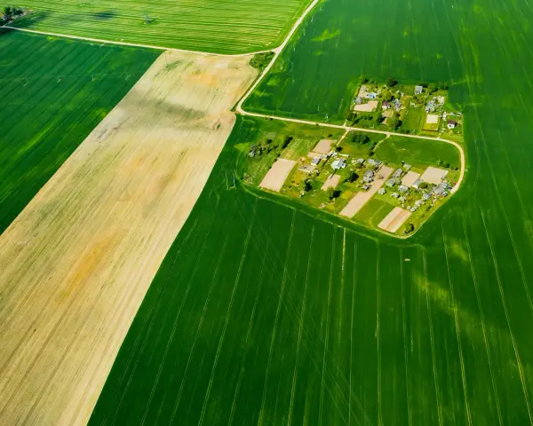 Widok z lotu ptaka na pola uprawne i wieś. Różne odcienie zieleni i żółcieni sugerują różne rodzaje gleby, co może pomóc w zrozumieniu, jak sprawdzić klasę gruntu.