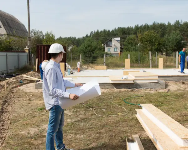 Inżynier z planami analizuje teren budowy, zastanawiając się, ile arów pod dom będzie potrzebne. Pracownicy w niebieskich kombinezonach kontynuują montaż ścian.