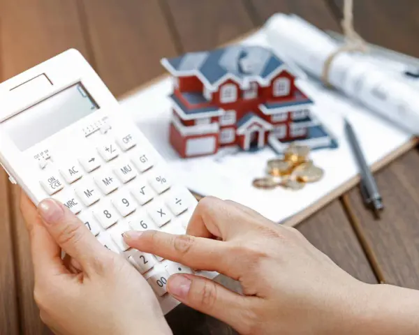 Dłoń na kalkulatorze, obok model domu, monety i plany. Wycena działki przez rzeczoznawcę będzie kluczowa dla banku od marca 2026.