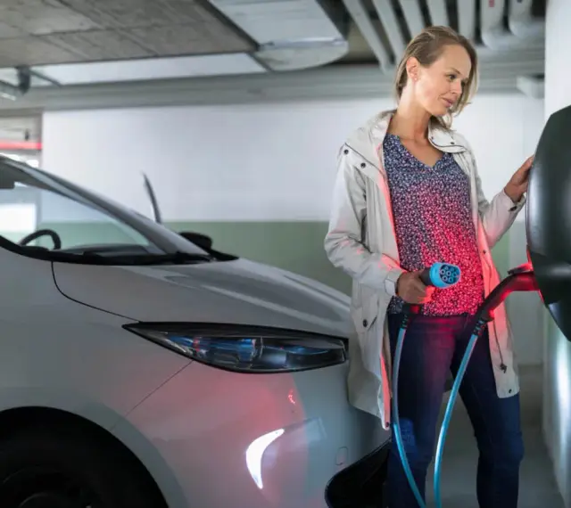 Kobieta ładuje biały samochód elektryczny. Czy od 2026 roku każda działka będzie musiała mieć przyłącze do ładowania aut elektrycznych?