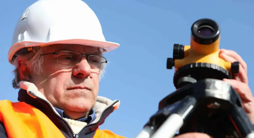 Geodeta w kasku i kamizelce odblaskowej pracuje z niwelatorem. Zastanawiasz się, ile kosztuje geodeta przy budowie domu?