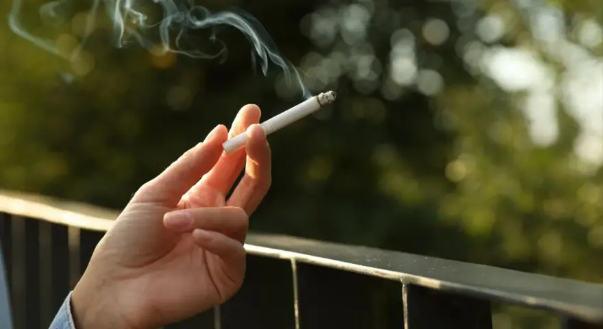 Dłoń trzyma zapalonego papierosa, z którego unosi się dym. Czy to prawda o rzekomym zakazie palenia papierosów na balkonach w nowych regulaminach?