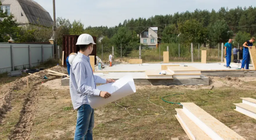 Inżynier z planami analizuje teren budowy, zastanawiając się, ile arów pod dom będzie potrzebne. Pracownicy w niebieskich kombinezonach kontynuują montaż ścian.