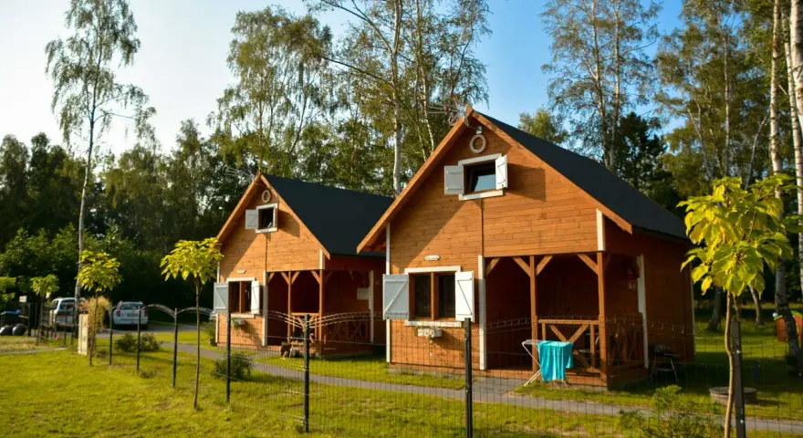 Dwa drewniane domki rekreacji indywidualnej, idealne na wynajem, stoją na zielonej trawie wśród drzew.
