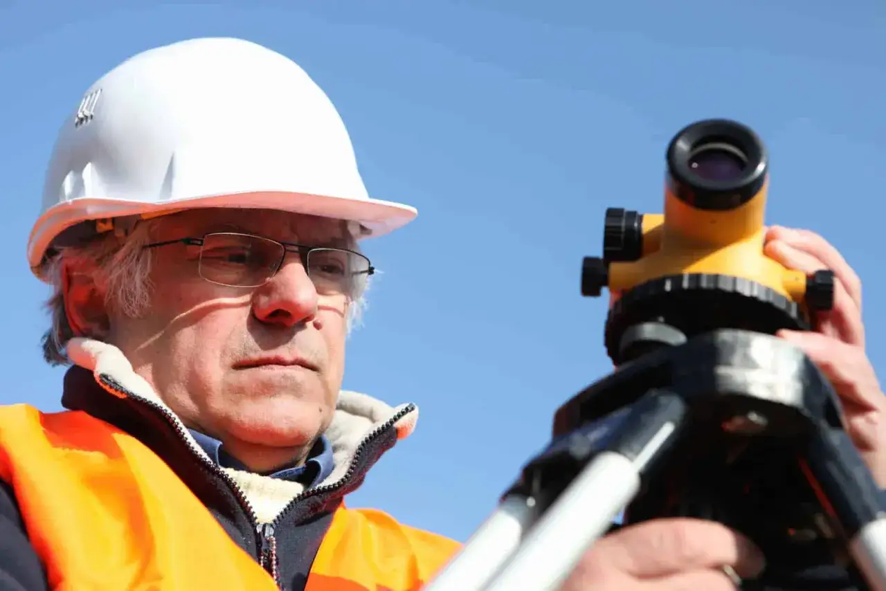 Geodeta w kasku i kamizelce odblaskowej pracuje z niwelatorem. Dowiedz się, ile kosztuje geodeta przy budowie domu.